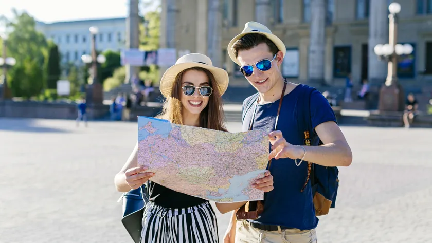 счастливая пара туристов в шляпах и солнцезащитных очках изучает бумажную карту города на залитой солнцем площади.