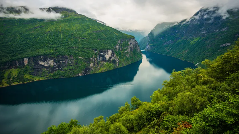 Вид сверху на извилистый норвежский фьорд с густой растительностью и облачным небом