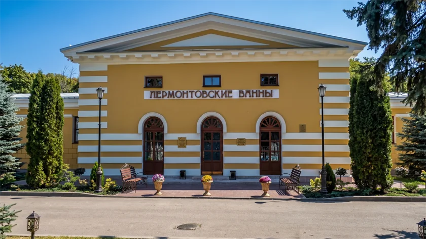 Исторический фасад отеля «Лермонтовские ванны» в Пятигорске. Горчично-желтое здание в классическом стиле с белой отделкой, арочными дверями и надписью на фасаде, окруженное зелеными туями и скамейками.